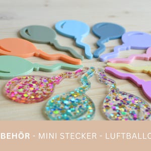 Pode incluir: Uma coleção de decorações coloridas em forma de balão. Cores pastel sólidas incluem laranja, verde, azul e roxo. Dois balões cheios de glitter também são visíveis. O texto na parte inferior diz "ZUBEHÖR - MINI STECKER - LUFTBALLON".