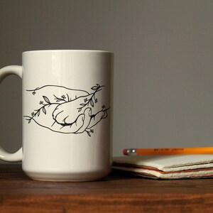 Pode incluir: Uma caneca de cer&acirc;mica branca com um desenho a tra&ccedil;o preto de duas m&atilde;os segurando-se, com detalhes florais. Um l&aacute;pis e um bloco de notas est&atilde;o sobre uma superf&iacute;cie de madeira. A caneca tem um tamanho padr&atilde;o.