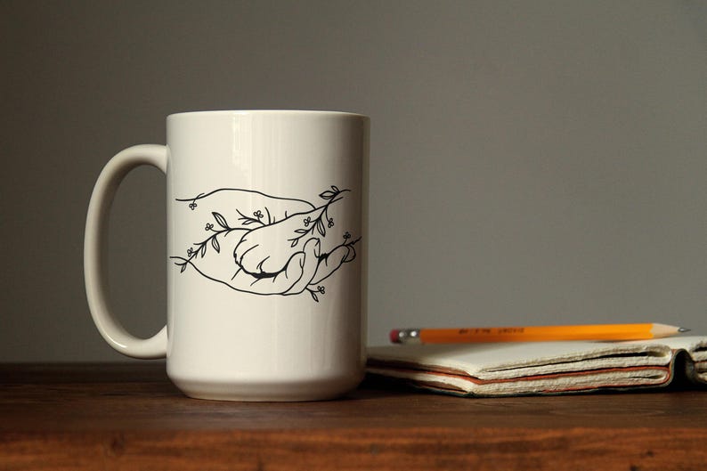 Pode incluir: Uma caneca de cer&acirc;mica branca com um desenho a tra&ccedil;o preto de duas m&atilde;os segurando-se, com detalhes florais. Um l&aacute;pis e um bloco de notas est&atilde;o sobre uma superf&iacute;cie de madeira. A caneca tem um tamanho padr&atilde;o.