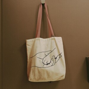 Pode incluir: Bolsa tote creme com um desenho a tra&ccedil;o preto de duas m&atilde;os. A bolsa tem al&ccedil;as rosa e est&aacute; pendurada em uma parede. A bolsa tote &eacute; feita de um material semelhante a lona.