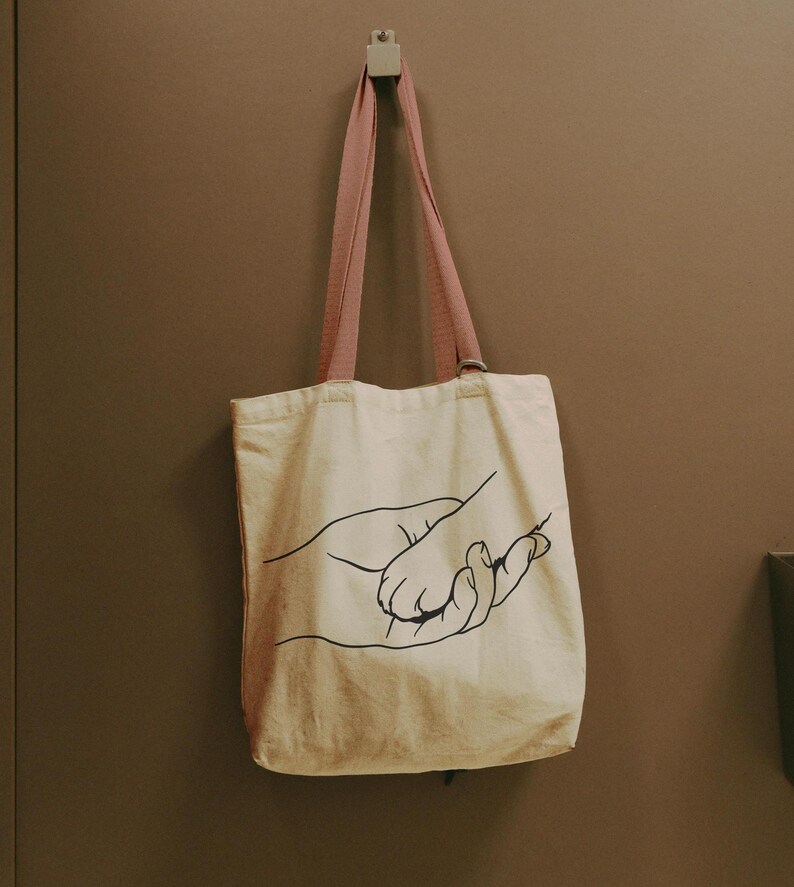 Pode incluir: Bolsa tote creme com um desenho a tra&ccedil;o preto de duas m&atilde;os. A bolsa tem al&ccedil;as rosa e est&aacute; pendurada em uma parede. A bolsa tote &eacute; feita de um material semelhante a lona.