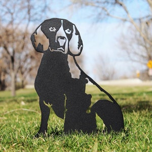 May include: Black metal silhouette of a beagle dog sitting in a grassy area. The dog is facing forward with its head tilted slightly to the side.