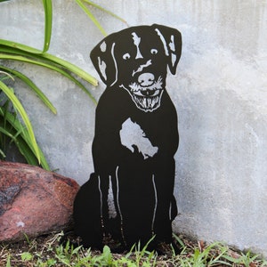 May include: Black metal silhouette of a dog sitting with its paws crossed. The dog has a playful expression and is cut out of metal.