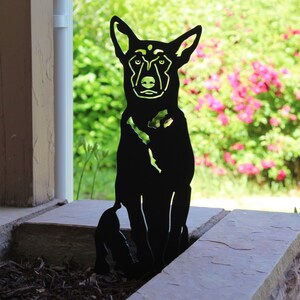 May include: Black metal silhouette of a dog sitting with its head tilted slightly to the side. The silhouette is placed in front of a bed of mulch and a stone wall.