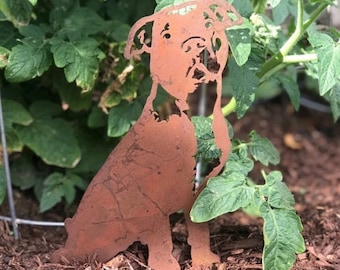 May include: A rusty metal silhouette of a dog sitting in a garden bed with brown mulch. The dog is facing forward with its head tilted slightly to the side. The dog's ears are perked up and its tail is curled.