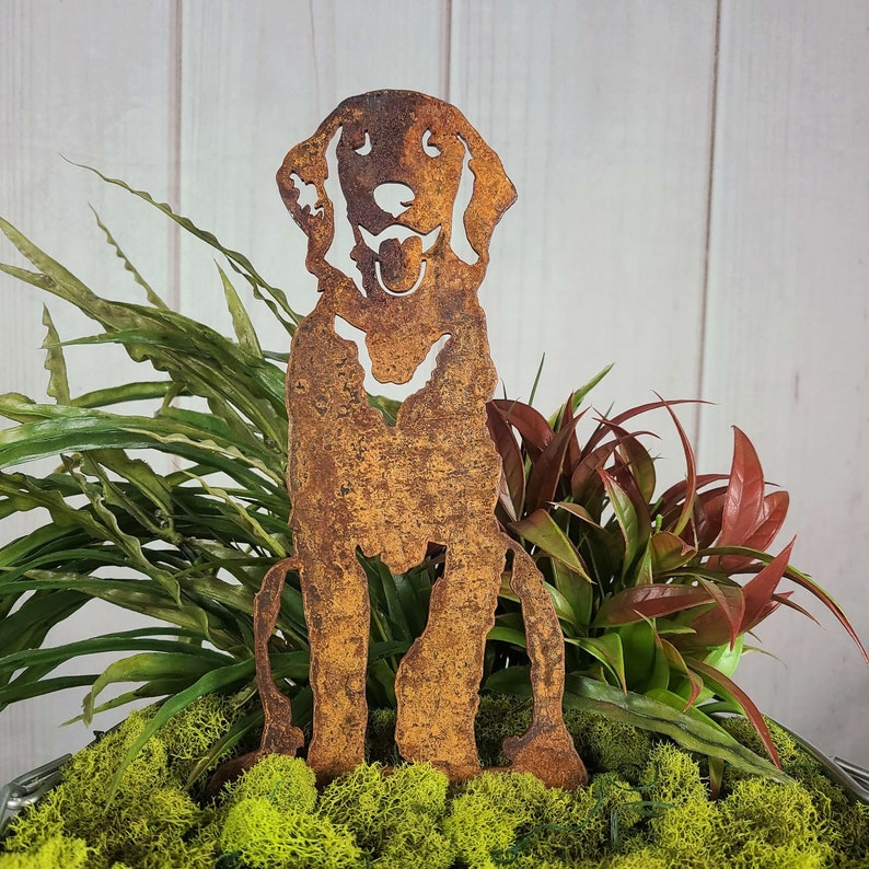 K&ouml;nnte beinhalten: Eine rostige Metallsilhouette eines sitzenden Hundes mit herausgestreckter Zunge. Die Silhouette ist in einem Topf mit gr&uuml;nem Moos und Pflanzen platziert.