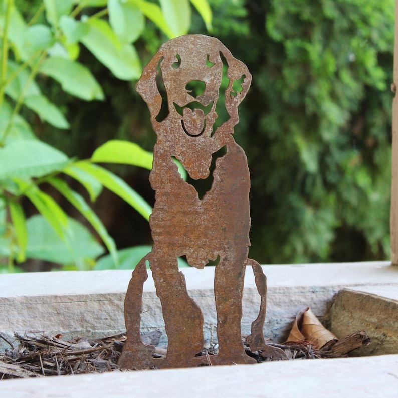 K&ouml;nnte beinhalten: Eine rostige Metallsilhouette eines Hundes mit einem fr&ouml;hlichen Ausdruck, die auf einem Bett aus braunem Mulch steht.