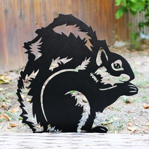 May include: Black metal silhouette of a squirrel, standing on a wooden surface. The squirrel is facing right and has a detailed design with fur and a bushy tail.