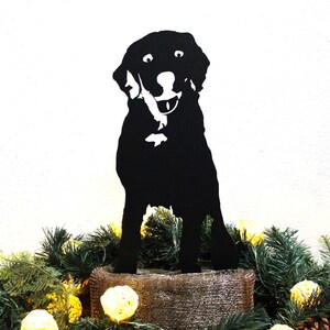 May include: Black metal silhouette of a dog sitting on a brown burlap base with a green garland of pine branches and white lights.