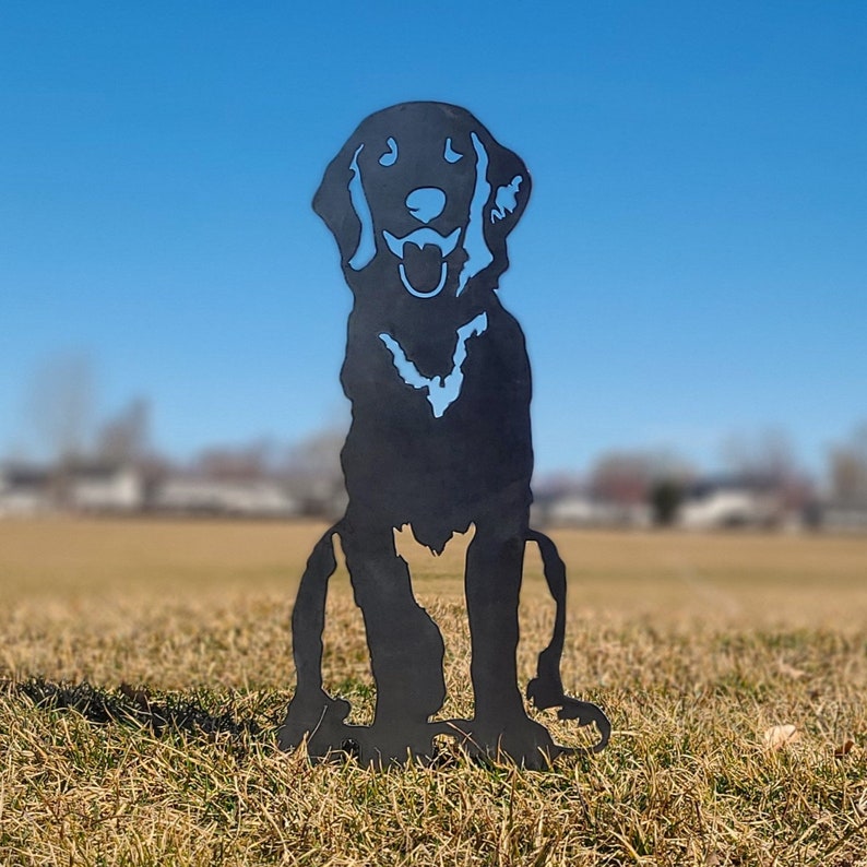 K&ouml;nnte beinhalten: Schwarze Metallsilhouette eines sitzenden Hundes mit offenem Maul, als w&uuml;rde er bellen. Die Silhouette ist auf einem grasbewachsenen Feld mit blauem Himmel im Hintergrund platziert.
