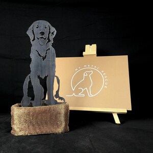 K&ouml;nnte beinhalten: Eine schwarze Metallsilhouette eines Hundes, der auf einem goldenen Drahtgeflecht-Sockel sitzt. Der Hund sitzt mit leicht geneigtem Kopf. Der Sockel ist rechteckig und aus feinem Drahtgeflecht gefertigt. Der Hintergrund ist schwarz. Das Bild zeigt auch ein Schild mit dem Text "My Metal Rescue".