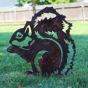 May include: A rusty metal squirrel silhouette with a detailed design. The squirrel is facing left and has a bushy tail. The silhouette is standing on a green grassy surface.