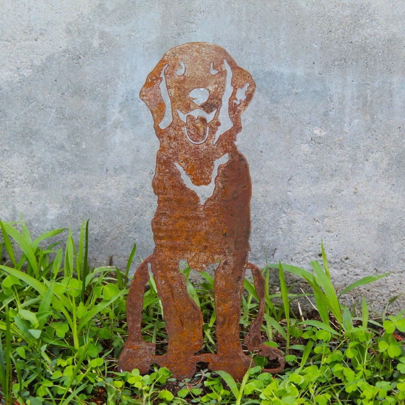 K&ouml;nnte beinhalten: Eine rostige Metallsilhouette eines sitzenden Hundes mit herausgestreckter Zunge. Die Silhouette ist vor einer grauen Betonwand und gr&uuml;nem Gras platziert.