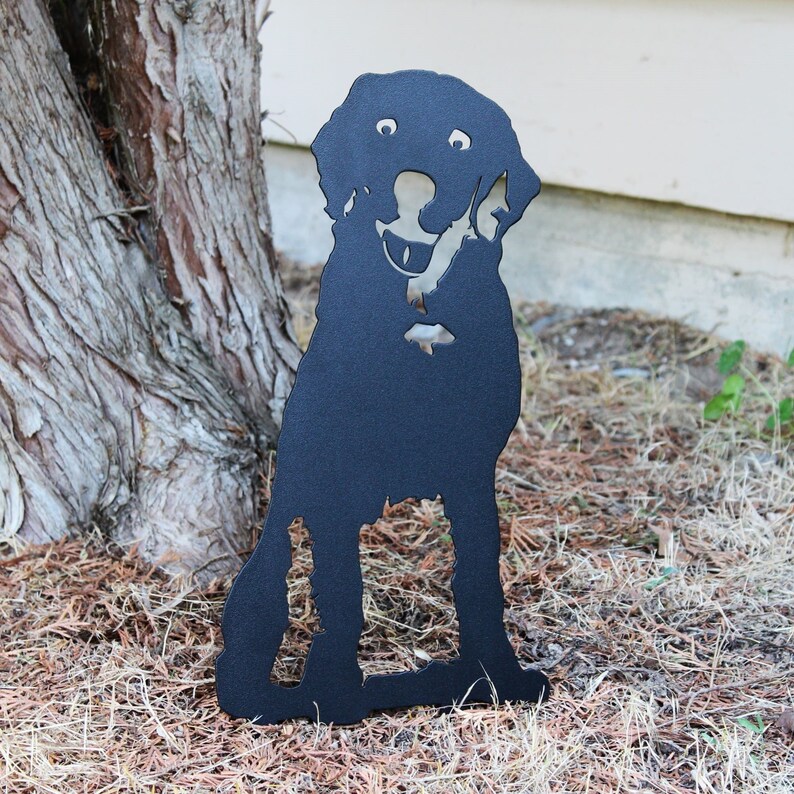 May include: Black metal silhouette of a dog sitting with its head tilted and mouth open, as if it is holding something in its mouth. The silhouette is placed on a bed of brown pine needles.