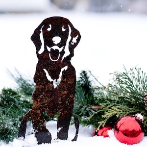 K&ouml;nnte beinhalten: Eine rostige Metallsilhouette eines Hundes, der im Schnee sitzt, mit immergr&uuml;nen Zweigen und roten Ornamenten drumherum.