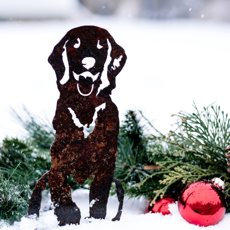 K&ouml;nnte beinhalten: Eine rostige Metallsilhouette eines Hundes, der im Schnee sitzt, mit immergr&uuml;nen Zweigen und roten Ornamenten drumherum.