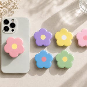 Pode incluir: Um smartphone cinza claro com um suporte de telefone em forma de flor rosa. Seis suportes de telefone coloridos em forma de flor em roxo, amarelo, rosa, azul e verde também são exibidos. As pegas têm um centro amarelo ou rosa.