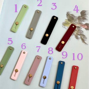 Puede incluir: Una colección de diez agarres rectangulares para teléfono en varios colores, incluyendo verde, gris, negro, burdeos, rosa, lavanda, azul y rojo. Cada agarre presenta un botón circular dorado y un pequeño orificio para el soporte de los dedos.