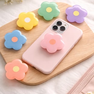 May include: A pink smartphone with a flower-shaped phone grip attached, alongside six flower-shaped grips in yellow, green, purple, blue, and orange. The grips have a contrasting centre colour and are arranged on a wooden surface.