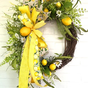 May include: A lemon-themed wreath with a yellow and white ribbon. The wreath is made of grapevine and is decorated with artificial lemons, leaves, and flowers.