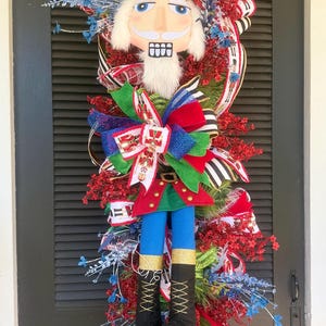 May include: A festive Christmas wreath featuring a plush nutcracker soldier. The wreath has a red and gold hat, blue pants, and black boots. It is decorated with red berries, blue flowers, and colorful ribbons.