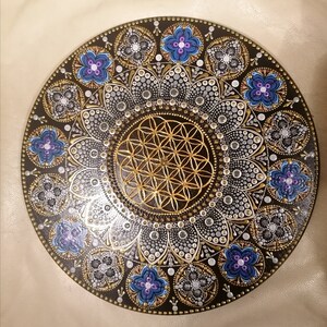 May include: A circular mandala painted on a wooden surface. The mandala features a flower of life design in gold, surrounded by a pattern of silver and gold dots. The outer edge of the mandala is decorated with blue and purple flowers.