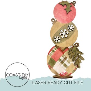 May include: A wooden Christmas ornament stack with three round ornaments. The top ornament is pink with a holly leaf design. The middle ornament is tan with snowflake accents. The bottom ornament has a plaid pattern. The text "LASER READY CUT FILE" is at the bottom.