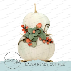 Puede incluir: Una decoración de calabaza apilada blanca con un grupo de hojas verdes, bayas naranjas y una bellota. El diseño incluye un tallo en la parte superior y un logotipo circular con el texto COAST DIY DIGITAL y LASER READY CUT FILE.