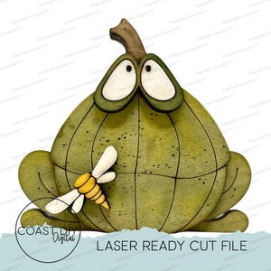 May include: A flat wooden cutout of a green pumpkin-shaped frog with large white eyes, a brown stem, and a small yellow and white bee attached to its front. The design features black etched lines and speckles on the green surface.