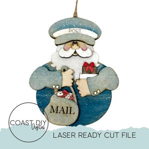 Op de afbeelding: Een houten kerstversiering van de kerstman met een blauwe postbodehoed met de tekst "POST". De versiering heeft een witte baard, roze wangen en houdt een postzak met het opschrift "MAIL" vast. Een rode strik siert een klein cadeau. De woorden "COAST DIY Digital" en "LASER READY CUT FILE" zijn ook zichtbaar.