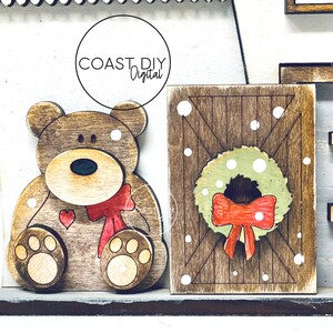 May include: Wooden Christmas decor with a brown bear wearing a red bow and a wooden sign with a green wreath and red bow. The sign has a wood grain pattern and white dots.