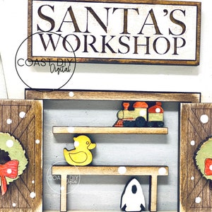 May include: A wooden sign with the text "Santa's Workshop" in white lettering on a brown background. The sign is decorated with white snowflakes. The sign is part of a larger wooden craft project that includes a toy train, a yellow rubber duck, and a white rocket.