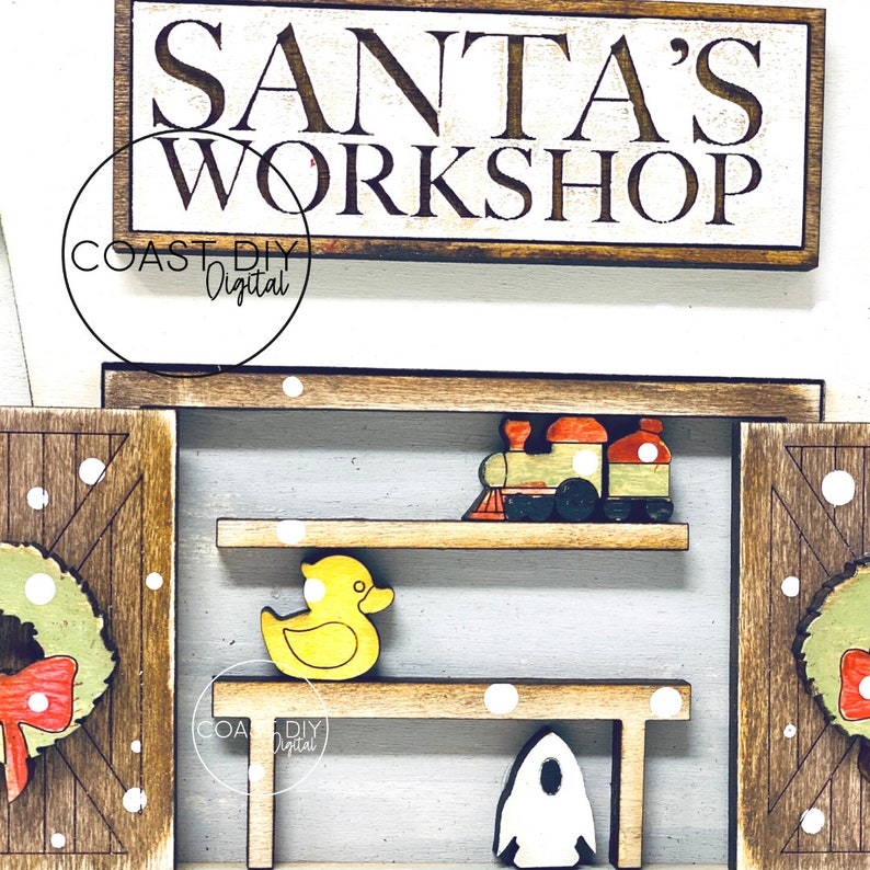 May include: A wooden sign with the text "Santa's Workshop" in white lettering on a brown background. The sign is decorated with white snowflakes. The sign is part of a larger wooden craft project that includes a toy train, a yellow rubber duck, and a white rocket.