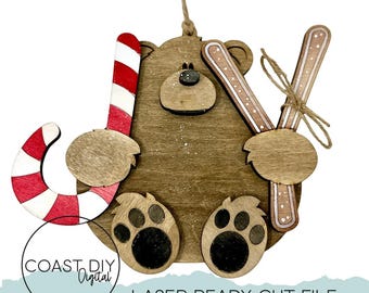 May include: Wooden bear Christmas ornament with a candy cane and skis. The bear has paw prints and a small nose. The candy cane is red and white striped. The skis are brown with white and pink accents. The ornament is laser-ready cut file.
