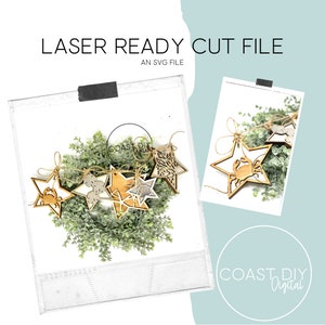 Puede incluir: Un conjunto de cinco estrellas de madera cortadas con láser con varios temas náuticos, incluyendo un cangrejo, conchas marinas y una estrella de mar. Los adornos están enhebrados con un cordel y se muestran en una corona verde. La imagen incluye el texto "COAST DIY Digital".