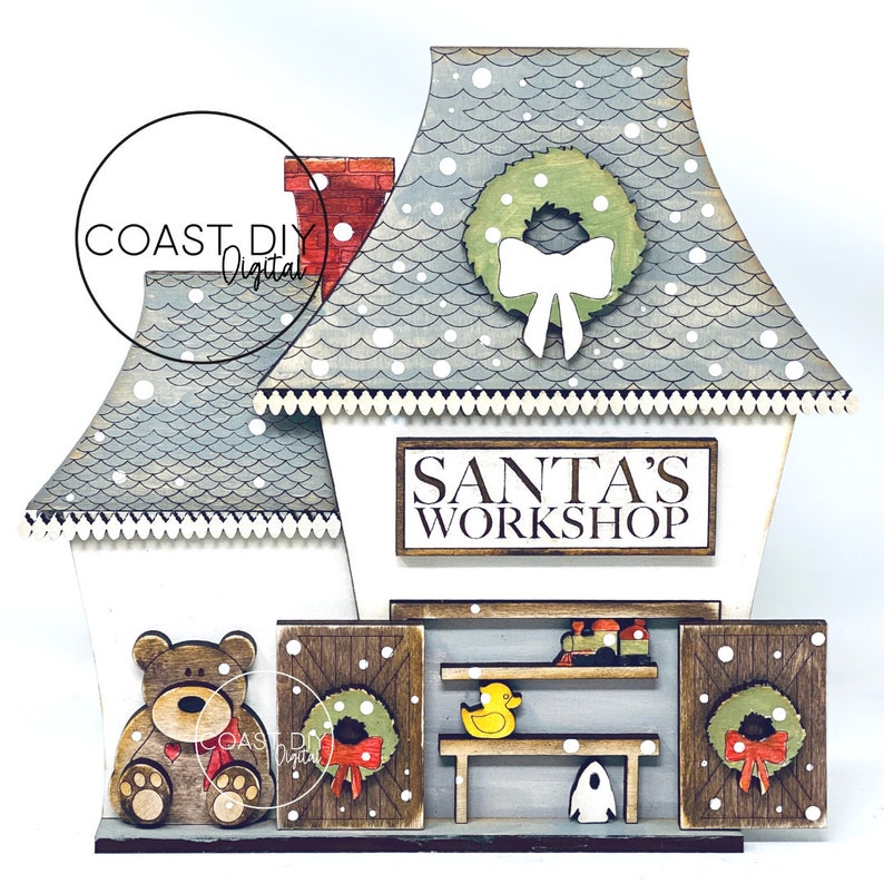 May include: A wooden Santa's Workshop sign with a white background and brown lettering. The sign is attached to a white building with a blue roof and a green wreath with a white bow. The building has two doors with wreaths and a teddy bear standing in front of the building.