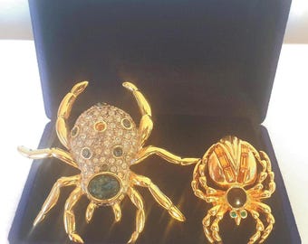 May include: Two gold-coloured spider brooches with gemstone accents sit inside a dark blue velvet box. The larger spider features a body encrusted with clear and coloured crystals, while the smaller one has a black centre stone.