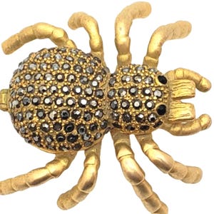 May include: A gold-toned spider brooch with a textured finish. The spider's body is covered in small, dark gray gemstones. The legs and other details are in a matte gold color, creating a striking contrast. The brooch is designed with intricate details.