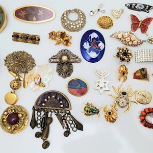 May include: A selection of vintage brooches and pins in diverse shapes and designs. Features a jeweled spider, a butterfly, and a floral brooch. The brooches are in gold, silver, and various colours.