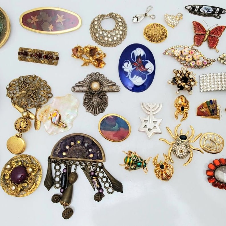 May include: A selection of vintage brooches and pins in diverse shapes and designs. Features a jeweled spider, a butterfly, and a floral brooch. The brooches are in gold, silver, and various colours.