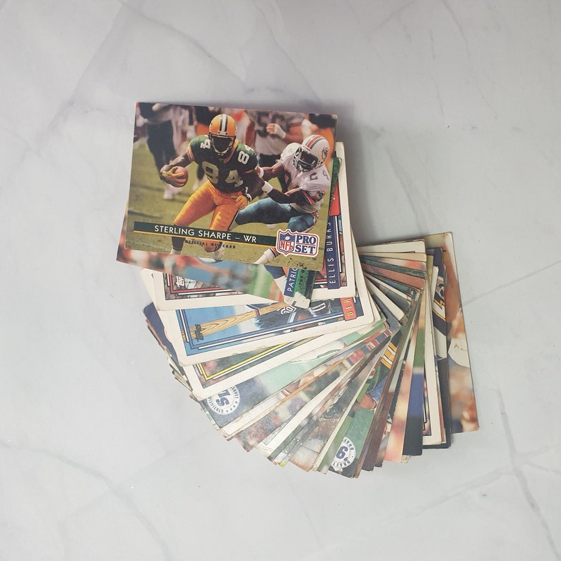 May include: A stack of sports trading cards featuring a variety of players and teams. The top card features a football player in a green uniform with the text "STERLING SHARPE - WR" and "PRO SET" visible.
