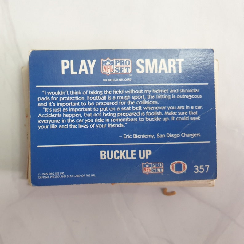 May include: A blue and white card with the text "Play Smart" and "Buckle Up". The card features a quote from Eric Bieniemy, a former San Diego Chargers player, about the importance of wearing a helmet and seatbelt for safety. The card is from the 1992 Pro Set NFL card collection.