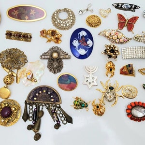 May include: A collection of vintage brooches and pins featuring a variety of designs, including floral, geometric, animal, and religious motifs. The brooches are made of metal, glass, and other materials and are decorated with rhinestones, pearls, and other embellishments.