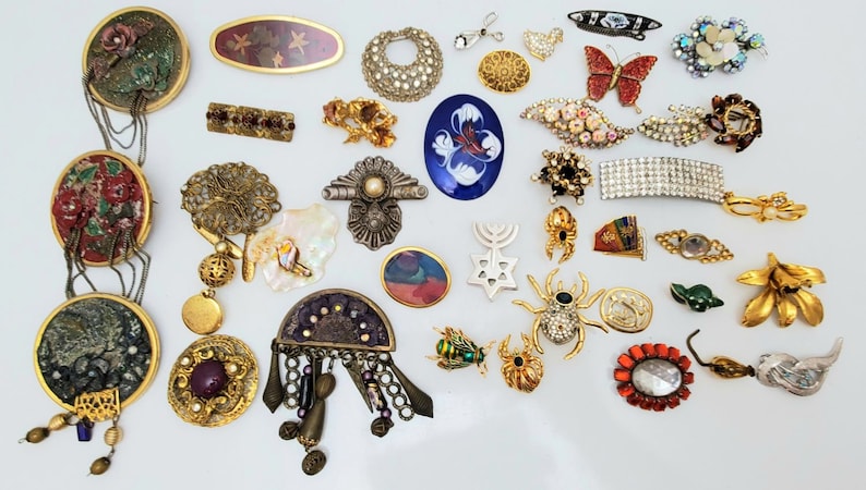 May include: A collection of vintage brooches and pins featuring a variety of designs, including floral, geometric, animal, and religious motifs. The brooches are made of metal, glass, and other materials and are decorated with rhinestones, pearls, and other embellishments.