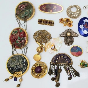 May include: A collection of assorted metal brooches and pins featuring various designs, including circular pendants with floral motifs, filigree patterns, enamel accents, dangling beads, chains, and geometric shapes in gold, silver, and multi-coloured finishes.