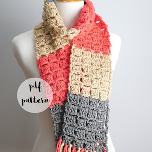 Può includere: Una sciarpa all'uncinetto con un motivo di colore grigio, corallo e beige. La sciarpa ha una frangia sul fondo ed è esposta su un manichino bianco. Il testo "pdf pattern" è visibile nell'immagine.