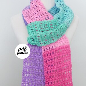 Peut inclure: Une écharpe au crochet multicolore dans les tons de violet, rose et turquoise. L'écharpe est présentée sur un mannequin et présente un motif ajouré. Les textes "pdf pattern" et "fiber flux" sont visibles.