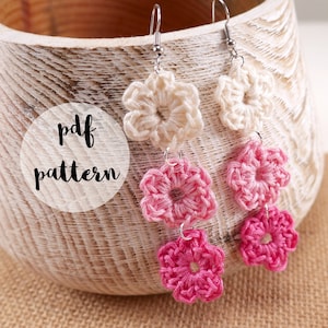 May include: Handmade crochet flower earrings in cream and pink. The earrings are displayed in a wooden bowl. The text "pdf pattern" is visible on the left side of the image. The earrings are a unique accessory.