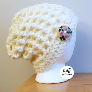 May include: Cream-colored crochet hat with a decorative button featuring a floral design. The hat has an open weave pattern and is displayed on a white mannequin head. The image includes the text "pdf pattern".