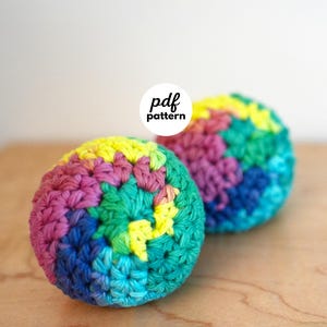 May include: Two spherical crochet balls featuring a vibrant, multi-coloured yarn pattern in shades of yellow, pink, teal, and blue. A circular white label with the black text pdf pattern is positioned between the two spheres.